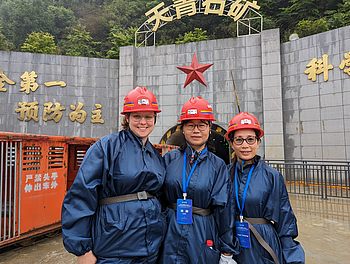 Drei Frauen mit Helm vor einem Stollenmundloch. Neben und über dem Mundloch sind ein Stern und chinesische Schriftzeichen zu sehen